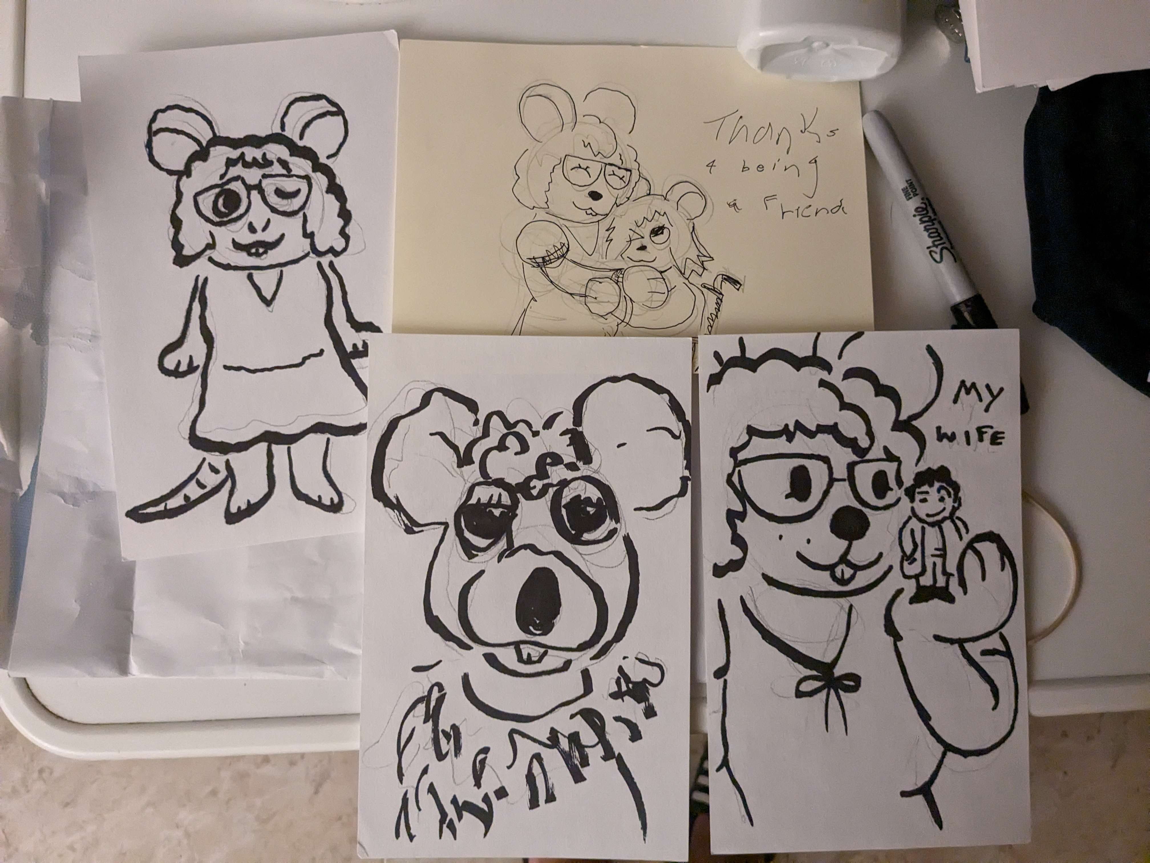 four drawings on a table. one is of katie as a little mouse wearing a dress with curly hair. another is of katie the mouse hugging marie the mouse, a smaller mouse with straight short hair in a wheelchair. text reads 'thanks 4 being a friend'. another drawing is of katie the mouse holding a small columbo from the tv series columbo. he is a small man in a trenchcoat. text has him saying 'my wife'. the final drawing is of a rodent-like puppet decorated with ruffles around her collar.